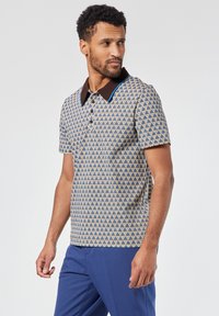 Lyhythihainen poolopaita, jossa on kuviollinen kangas, jossa on sinisen, keltaisen ja ruskean värejä; kaulus on yksivärinen ruskea sinisillä korostuksilla.