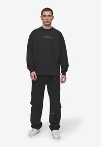 Mustat pitkät hihat sweatshirt, jossa on valkoinen logo rinnassa, yhdistettynä mustiin cargo-housuihin, joissa on sivutaskut, ja valkoisiin lenkkareihin.
