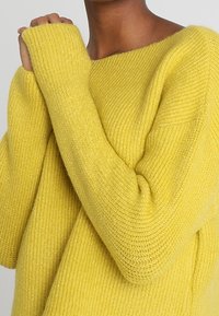 Osoba oblečená v měkkém žlutém pleteném svetr s dlouhými rukávy, ruce složené u klíční kosti, neutrální pozadí.