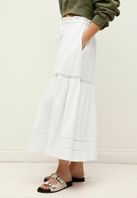 Jupe maxi blanche en coton avec un design à volants et un ourlet en dentelle. Associée à des sandales beiges avec des semelles noires et deux sangles ajustables.