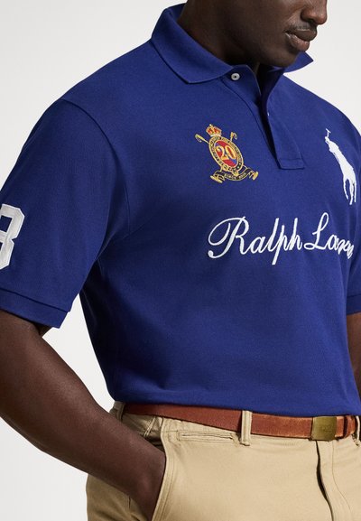 Mann iført en kongelig blå Ralph Lauren polo-skjorte med broderte logoer, beige bukser og et brunt belte, med hånden i lommen.