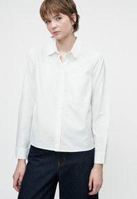 Camicia bianca con bottone, maniche lunghe, taschino singolo sul petto, tessuto leggero con una trama liscia, abbinata a pantaloni in denim scuro.