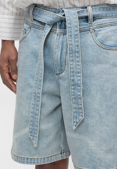 Ανοιχτό μπλε denim σορτς με ζώνη από το ίδιο ύφασμα δεμένη στη μέση, φορεμένα με ριγέ πουκάμισο μακριών μανικιών.