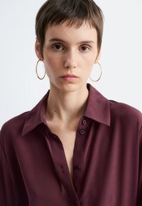 Burgundy blouse with a collar, featuring a button placket and soft, smooth texture. Model wears large gold hoop earrings.
