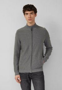 Grijze sweater met rits en hoge kraag, geribbelde manchetten en zoom. Voorzien van zijzakken en een gestructureerd patroon. Draagt over een donkere spijkerbroek.