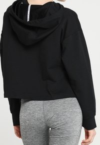 Felpa nera corta con cappuccio regolabile; presenta una vestibilità morbida e un tessuto di texture soffice. Abbinata a leggings grigi.