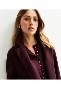 Cappotto bordeaux con colletto alto, sopra un top testurizzato con bottoni dorati e scollatura rotonda. Lunghi capelli ondulati incorniciano il viso.