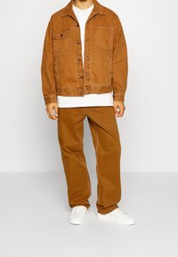 Veste en denim marron avec deux poches poitrine, boutons de fermeture et col, associée à un pantalon marron assorti et des baskets blanches.