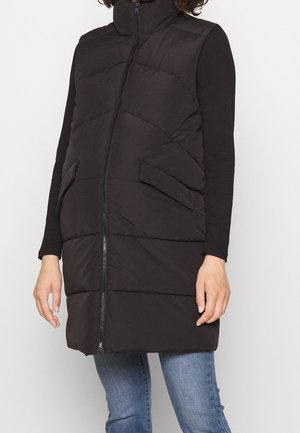 Sort quiltet ærmeløs vest med lynlås foran og lommer med klap, båret over en sort langærmet top og blå jeans.