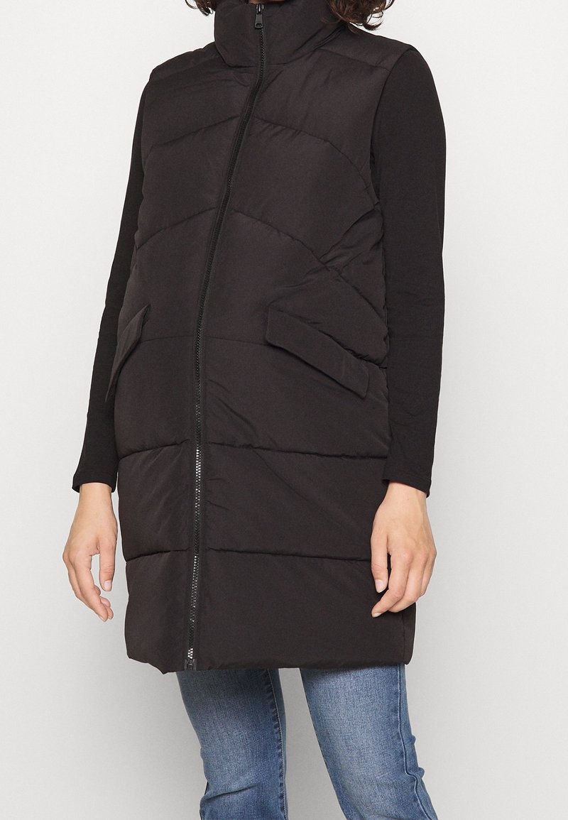 Veste sans manches matelassée noire avec fermeture éclair frontale et poches à rabat portée sur un haut à manches longues noir et un jean bleu.
