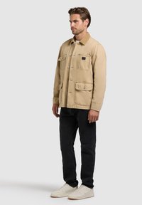 Homme portant une veste beige à boutons avec quatre poches, un pantalon noir et des baskets blanches, debout devant un fond clair uni.