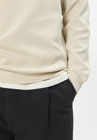 Béžový dlhý rukáv s okrúhlym výstrihom a jemnou textúrou, vrstvený ponad biely lem. Nosený s čiernymi plisovanými nohavicami.