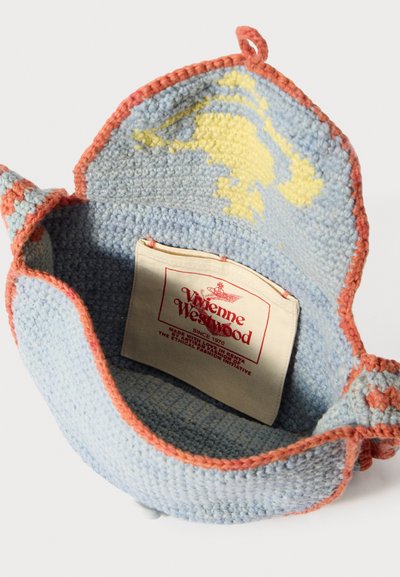 Tamborētas soma ar gaiši zilu korpusu un korāla krāsas apmale, tajā redzams dzeltens raksts. Iekšpusē ir etiķete ar zīmola informāciju.