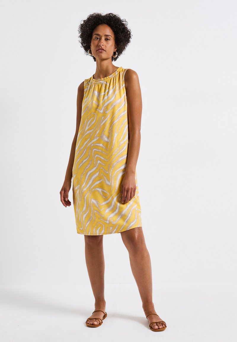 Ärmelloses gelbes Kleid mit weißen wellenförmigen Streifen, das einen gerafften Ausschnitt aufweist. Weicher Stoff mit lockerer Passform, der über die Knie reicht.
