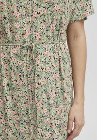 Robe florale verte avec des motifs roses et orange, présentant des manches courtes et une taille à cordon pour un ajustement réglable. Texture de tissu lisse.