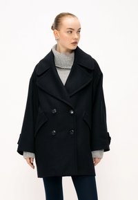 Cappotto doppio petto di lana nera con grandi revers, tasche laterali e maniche oversize, indossato su un dolcevita grigio chiaro a coste.