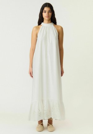 Woman with long dark hair wearing a sleeveless white halter neck maxi dress and beige espadrille shoes, standing against a plain background.