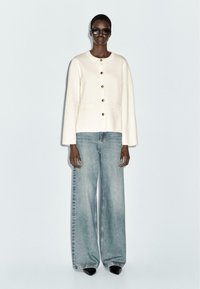 Crème-kleurige jas met gouden knopen, twee zakken, gecombineerd met hoog-wijde lichtblauwe denim jeans en zwarte schoenen.
