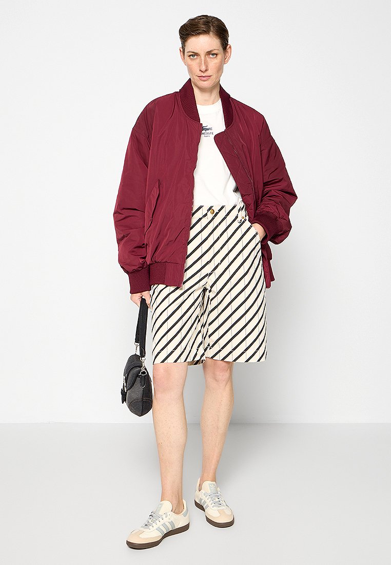 Bomber rouge sur un t-shirt blanc, associé à un short rayé beige. Porté avec des baskets beige et crème et un petit sac noir.