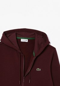 Felpa con zip color bordeaux, con accenti verdi, dotata di cappuccio regolabile e un piccolo logo ricamato sul lato sinistro del petto.