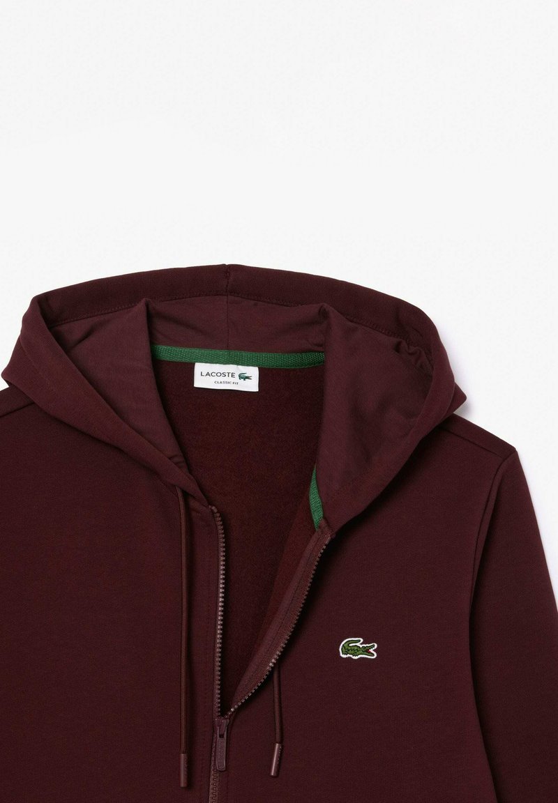 Hoodie zippé bordeaux avec des accents verts, doté d'une capuche à cordon et d'un petit logo brodé sur la poitrine gauche.