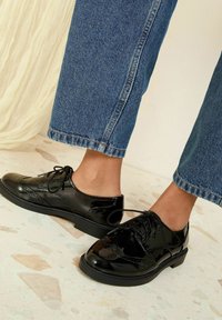 Brogues negre din piele lacuită, cu închidere cu șireturi, având design cu vârf în formă de aripă și perforații decorative, asociați cu blugi din denim albastru, scurți.