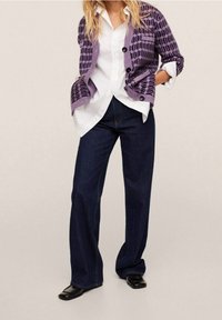 Žena v purpurovém vzorovaném kardiganu přes bílou halenku, tmavě modrých širokých kalhotách a černých botách se čtvercovou špičkou, stojí s rukama v kapsách.