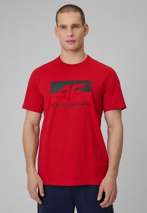 Mężczyzna w czerwonej koszulce z krótkim rękawem z czarnym logo "4F Sportswear Brand", stojący na jednolitym szarym tle.