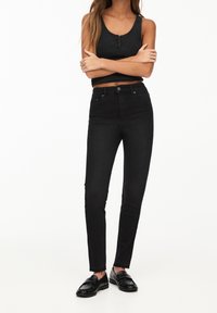 Jeans negros de cintura alta y ajuste slim, combinados con una blusa sin mangas negra. El conjunto presenta un hardware minimalista y una textura suave.