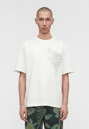 Άντρας με ξυρισμένο κεφάλι που φοράει λευκό μπλουζάκι με διακριτικό floral σχέδιο στο στήθος και σκούρο πράσινο παντελόνι με φυλλαράκια.