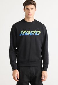 Young man wearing a black sweatshirt with blue and green gradient flame-style text "Hugo" on chest, standing against a plain white background.