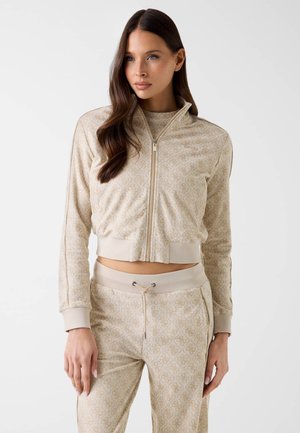 Femme aux longs cheveux bruns portant une veste beige à motifs zippée et un pantalon assorti à cordon, debout devant un fond clair et uni.