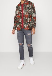 Heren overhemd met lange mouwen in een multikleurig camouflagepatroon in groen, rood en beige, gecombineerd met grijze distressed jeans en witte sneakers.