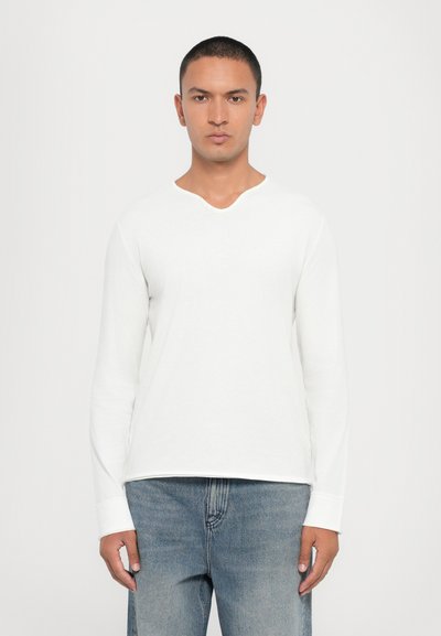 Maglietta bianca a maniche lunghe con leggero scollo a V, realizzata in cotone morbido. Presenta una vestibilità comoda e orlo cucito, abbinata a jeans azzurri chiari.