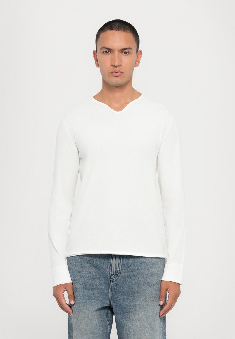 T-shirt blanc à manches longues avec un léger col en V, fabriqué en coton doux. Présente une coupe décontractée et un ourlet cousu, associé à un jean bleu clair.