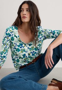 Haut à manches longues à motifs floraux avec un col rond, présentant des motifs verts et bleus sur un fond blanc, assorti à un jean foncé et des sandales marron.