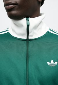 Green athletic jacket with white accents, high collar, and a zip front. Features three white stripes on shoulders and a small logo.