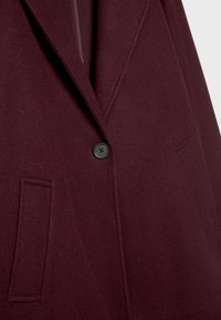 Burgunderfarbenen Wollmischmantel mit Reverskragen, einfachem schwarzen Knopfverschluss und zwei vorderen Paspeltaschen, mit einer weichen, strukturierten Oberfläche.