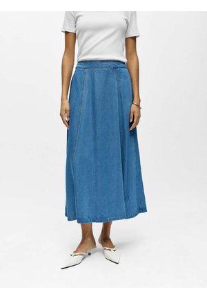 Frau trägt ein weißes kurzärmeliges Oberteil, einen blauen Denim-Midirock und weiße spitz zulaufende Slip-On-Schuhe, steht vor einem einfarbigen Hintergrund.