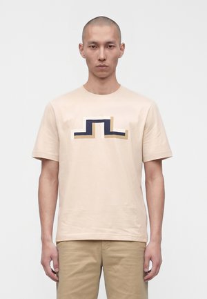 Homme à la tête rasée portant un t-shirt beige avec un motif géométrique bleu marine et beige, associé à un pantalon marron clair, debout devant un fond uni.