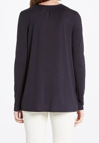 Maglietta a maniche lunghe di un blu navy scuro con scollo rotondo, dettaglio arricciato nella parte posteriore e tessuto morbido; abbinata a pantaloni di colore chiaro.