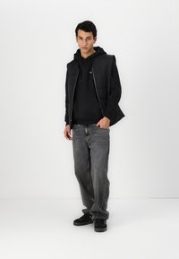 Giacca senza maniche nera sopra un hoodie nero, abbinata a jeans grigi. Il modello indossa sneakers nere, in piedi contro uno sfondo bianco.