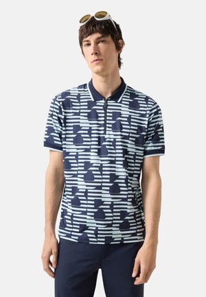 Jeune homme portant un polo à motifs géométriques bleu marine et blanc avec une fermeture éclair et des lunettes de soleil blanches sur la tête.