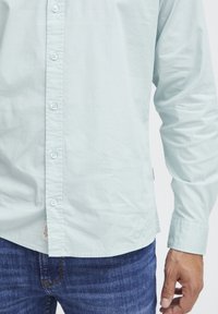 Camisa de botones azul claro hecha de tela suave, con dobladillo recto y mangas largas con puños abotonados. Se lleva con unos jeans de mezclilla oscuros.