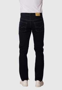 Jeans oscuros ajustados de mezclilla con costuras amarillas en contraste, cinco bolsillos y una etiqueta de cuero en la parte posterior de la cintura. Combinados con zapatillas blancas.