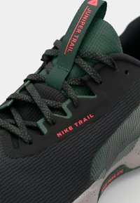 Baskets de trail Nike noires et vertes avec une tige texturée, un bout arrondi et un logo rouge. Elles sont équipées de lacets verts et d'une semelle grise à motifs.