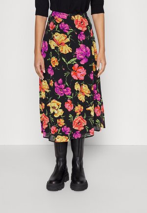 Frau trägt einen schwarzen Midi-Rock mit einem leuchtend pinken, orangefarbenen und gelben Blumenmuster, kombiniert mit schwarzen klobigen Stiefeln.