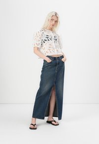 Top court en dentelle blanche avec des manches bouffantes sur une jupe maxi en denim bleu taille haute avec une fente frontale. Le modèle porte des tongs noires.
