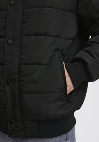 Sort puffet jakke med quiltet design, der har trykknapper, ribbede manchetter og sidelommer. Glat, isoleret tekstur.