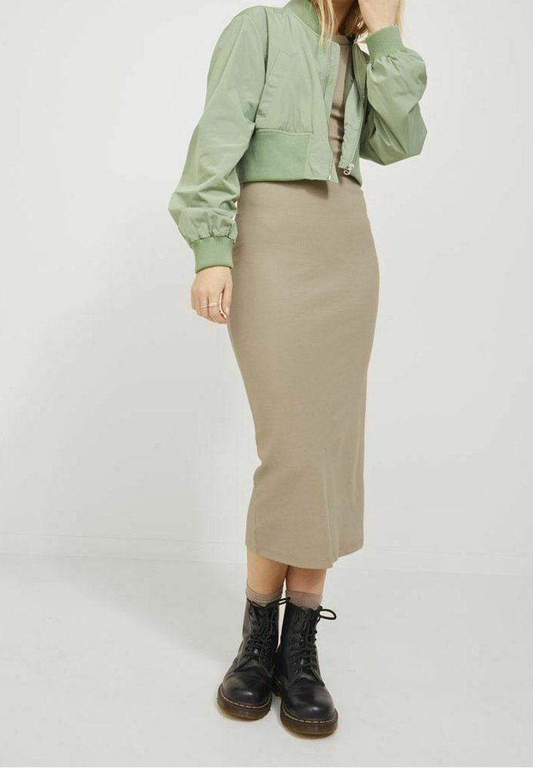 Femme portant une veste courte verte olive, une robe midi beige ajustée et des bottes de combat noires à lacets, debout devant un fond blanc.